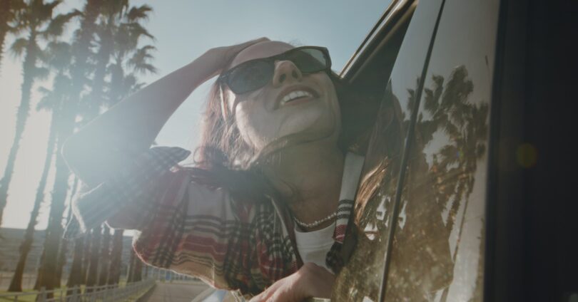 Smiling woman wearing sunglasses leans out of a car window as sunlight filters through a row of palm trees.