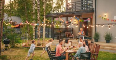 A family gathered together in the backyard of a large house. Kids are running around while adults are sitting.