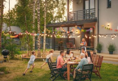 A family gathered together in the backyard of a large house. Kids are running around while adults are sitting.