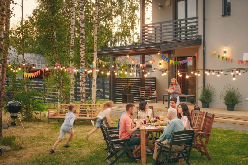 A family gathered together in the backyard of a large house. Kids are running around while adults are sitting.
