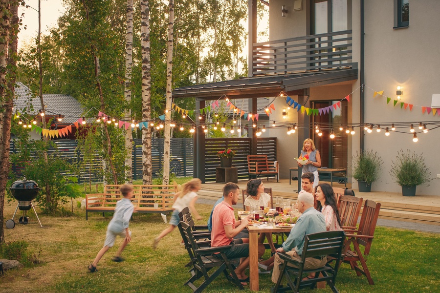 A family gathered together in the backyard of a large house. Kids are running around while adults are sitting.