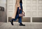 A woman walks in a camel coat, blue jeans, and black boots while carrying a structured brown handbag on a city street.