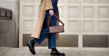 A woman walks in a camel coat, blue jeans, and black boots while carrying a structured brown handbag on a city street.