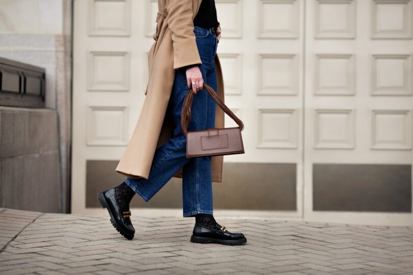 A woman walks in a camel coat, blue jeans, and black boots while carrying a structured brown handbag on a city street.