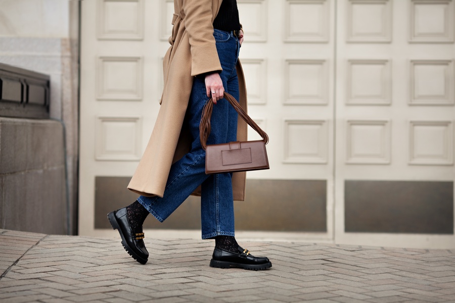 A woman walks in a camel coat, blue jeans, and black boots while carrying a structured brown handbag on a city street.