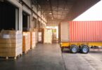 Pallets of packaged goods sit beside a freight container at a warehouse loading dock awaiting transportation.