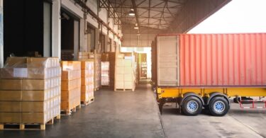 Pallets of packaged goods sit beside a freight container at a warehouse loading dock awaiting transportation.
