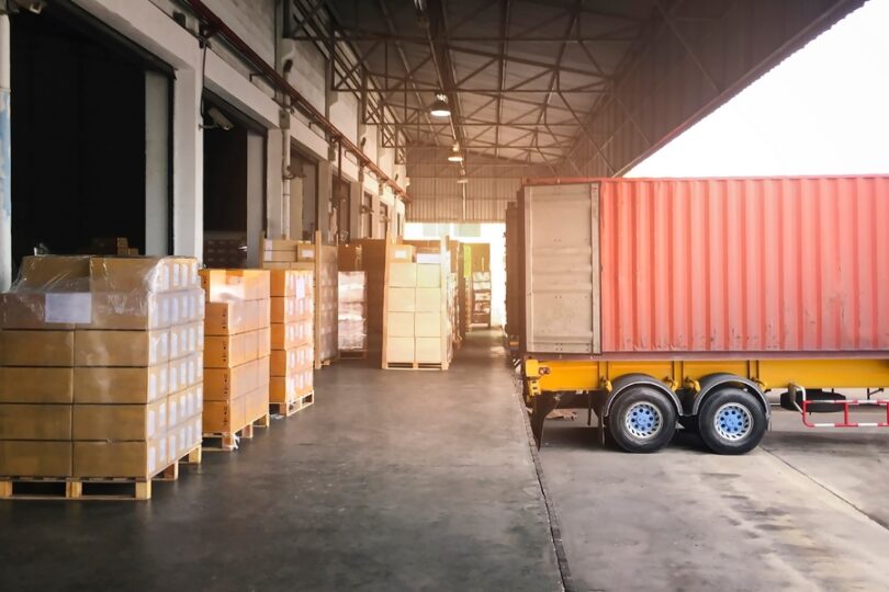 Pallets of packaged goods sit beside a freight container at a warehouse loading dock awaiting transportation.