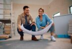 A man and a woman kneeling next to each other holding a large piece of paper inside a room undergoing a renovation.