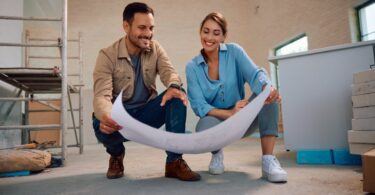 A man and a woman kneeling next to each other holding a large piece of paper inside a room undergoing a renovation.