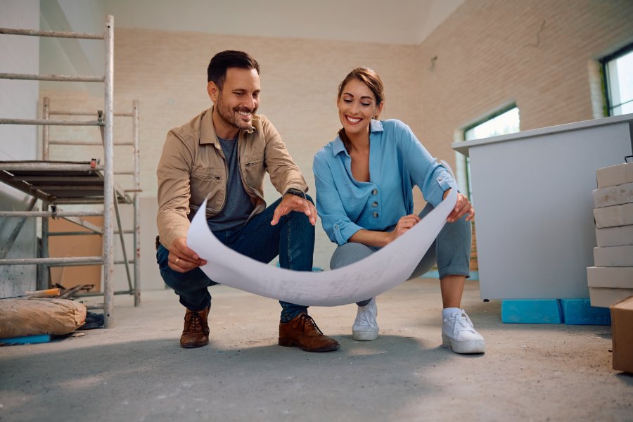 A man and a woman kneeling next to each other holding a large piece of paper inside a room undergoing a renovation.