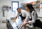 Two chefs cleaning a large, brightly lit commercial kitchen. One chef wipes a counter with a green rag.