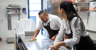 Two chefs cleaning a large, brightly lit commercial kitchen. One chef wipes a counter with a green rag.
