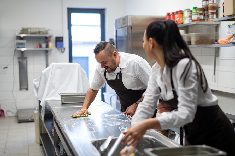 Two chefs cleaning a large, brightly lit commercial kitchen. One chef wipes a counter with a green rag.