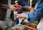 A vendor placing vegetables into a customer’s bag at a farmers market. Another vendor stands nearby holding products.