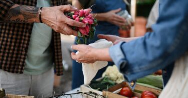 A vendor placing vegetables into a customer’s bag at a farmers market. Another vendor stands nearby holding products.