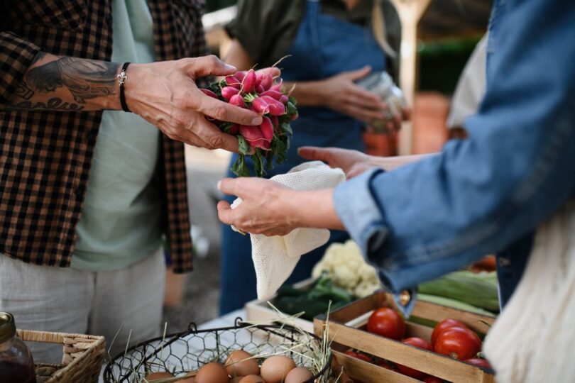 A vendor placing vegetables into a customer’s bag at a farmers market. Another vendor stands nearby holding products.