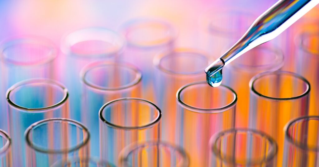A close-up of a collection of small lab test tubes with a dropper of liquid hovering over one test tube.
