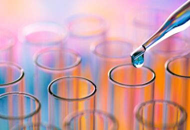 A close-up of a collection of small lab test tubes with a dropper of liquid hovering over one test tube.