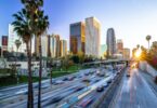 A downtown skyline behind a busy highway with cars, palm trees, and warm evening light in the sky above it.