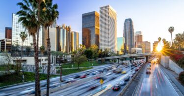 A downtown skyline behind a busy highway with cars, palm trees, and warm evening light in the sky above it.