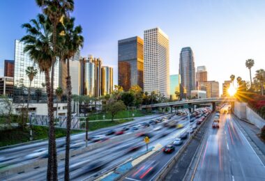 A downtown skyline behind a busy highway with cars, palm trees, and warm evening light in the sky above it.