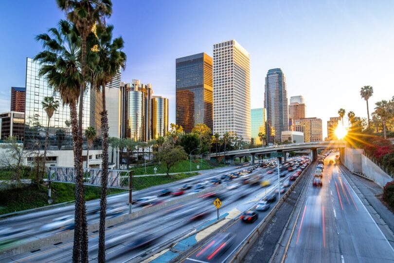 A downtown skyline behind a busy highway with cars, palm trees, and warm evening light in the sky above it.