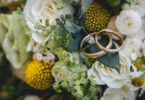 Two gold wedding bands rest on a floral bouquet of white roses, yellow blooms, small daisies, and green foliage