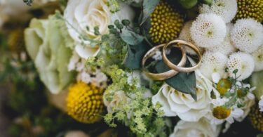 Two gold wedding bands rest on a floral bouquet of white roses, yellow blooms, small daisies, and green foliage