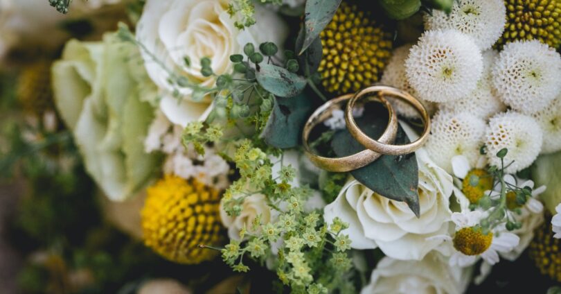 Two gold wedding bands rest on a floral bouquet of white roses, yellow blooms, small daisies, and green foliage