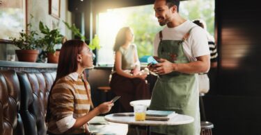 A woman sitting at a half-booth table holding a credit card while a man in a green apron with a card reader stands nearby.