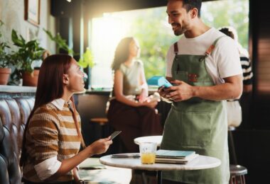 A woman sitting at a half-booth table holding a credit card while a man in a green apron with a card reader stands nearby.
